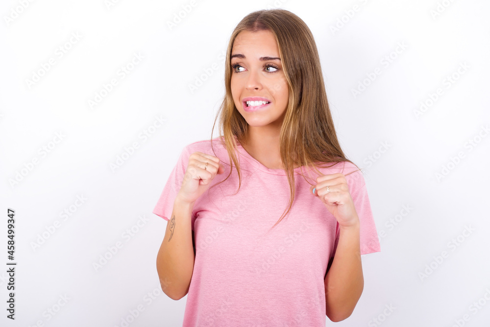 beautiful blonde girl wearing pink t-shirt on white background clenches fists and awaits for something nice happened looks away bites lips and waits announcement of results beautiful blonde girl wearing pink t-shirt on white background clenches fists and awaits for something nice happened looks away bites lips and waits announcement of results