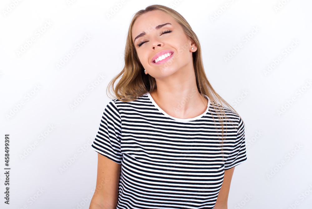 Positive beautiful blonde girl wearing striped t-shirt on white background with overjoyed expression closes eyes and laughs shows white perfect teeth Positive beautiful blonde girl wearing striped t-shirt on white background with overjoyed expression closes eyes and laughs shows white perfect teeth
