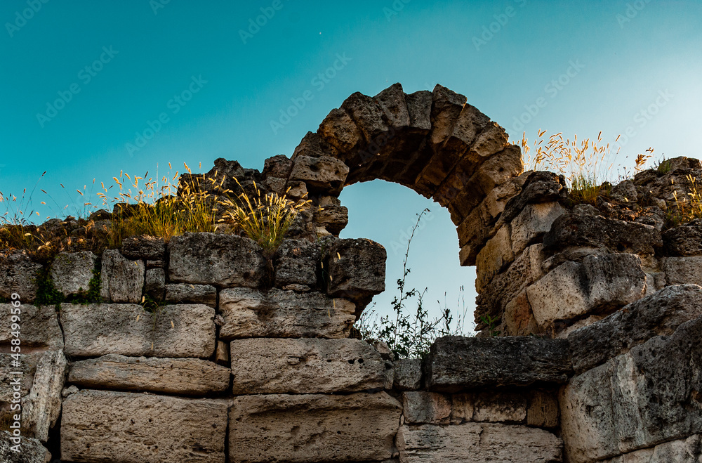 an arch of large stones on a high stone ancient dilapidated wall overgrown with grass against the blue sky