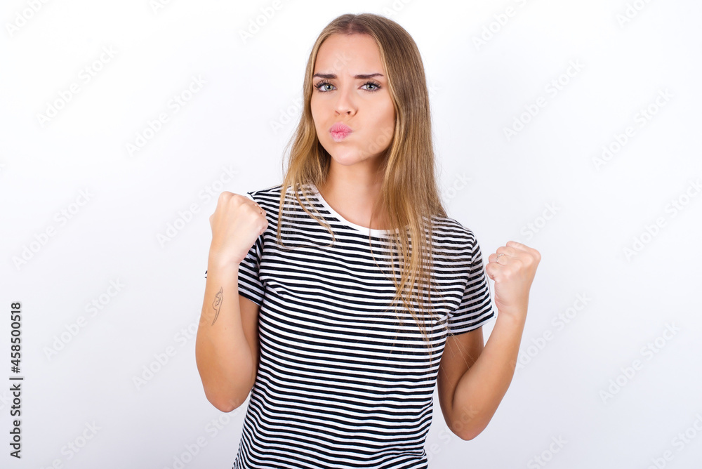 Fototapeta premium Irritated beautiful blonde girl wearing striped t-shirt on white background blows cheeks with anger and raises clenched fists expresses rage and aggressive emotions. Furious model