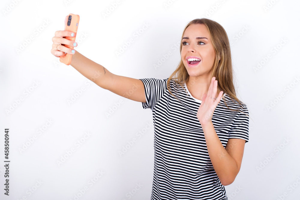 beautiful blonde girl wearing striped t-shirt on white background holds modern mobile phone and makes video call waves palm in hello gesture. People modern technology concept beautiful blonde girl wearing striped t-shirt on white background holds modern mobile phone and makes video call waves palm in hello gesture. People modern technology concept