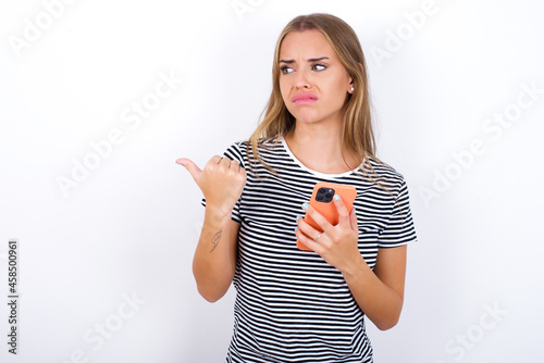 MObeautiful blonde girl wearing striped t-shirt on white background DEL points thumb away and shows blank space aside, holds mobile phone for sending text messages. photo