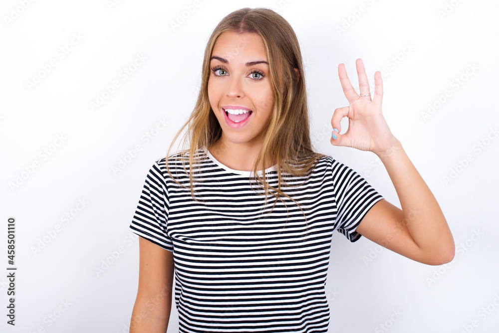 beautiful blonde girl wearing striped t-shirt on white background hold hand arm okey symbol toothy approve advising novelty news beautiful blonde girl wearing striped t-shirt on white background hold hand arm okey symbol toothy approve advising novelty news