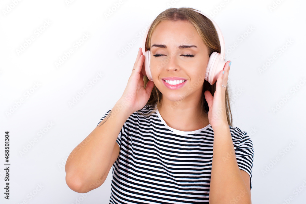 Pleased beautiful blonde girl wearing striped t-shirt on white background enjoys listening pleasant melody keeps hands on stereo headphones closes eyes. Spending free time with music Pleased beautiful blonde girl wearing striped t-shirt on white background enjoys listening pleasant melody keeps hands on stereo headphones closes eyes. Spending free time with music
