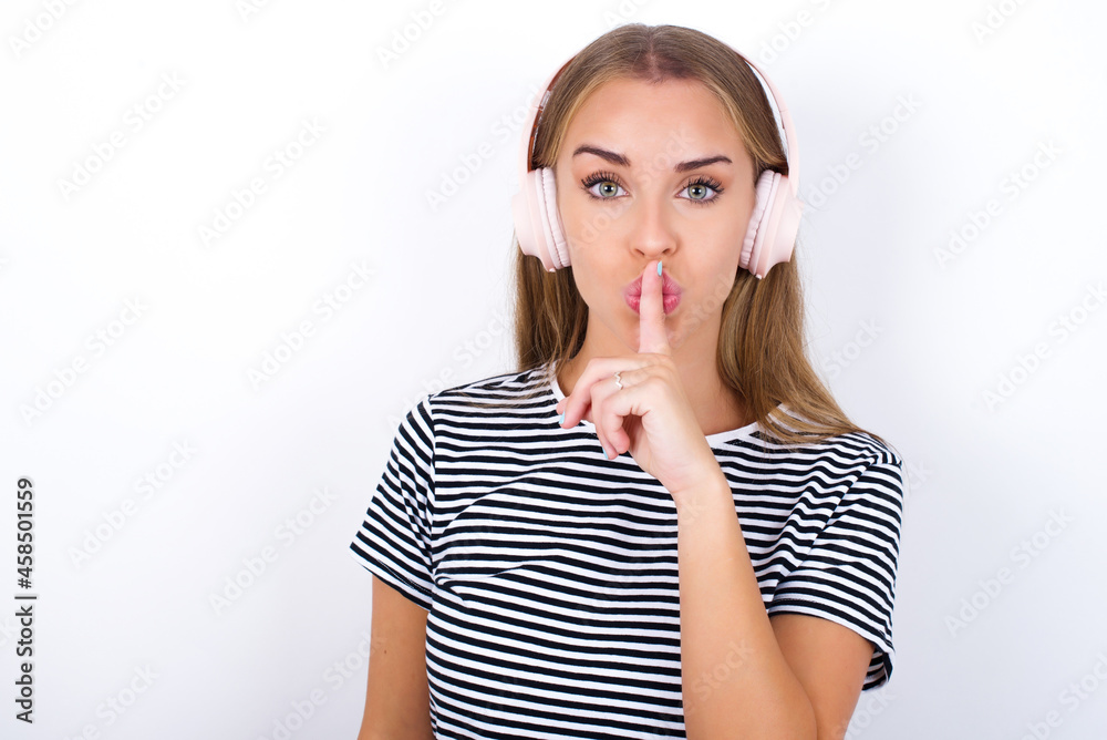 beautiful blonde girl wearing striped t-shirt on white background making hush gesture with finger on her lips wearing wireless headphones. Be quiet. beautiful blonde girl wearing striped t-shirt on white background making hush gesture with finger on her lips wearing wireless headphones. Be quiet.