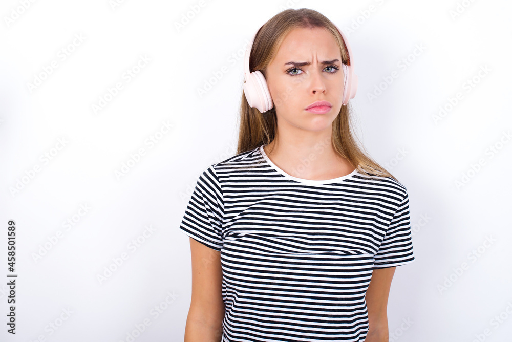 Serious displeased beautiful blonde girl wearing striped t-shirt on white background looks puzzled at camera being angry wears stereo headphones listens music while walking at street Serious displeased beautiful blonde girl wearing striped t-shirt on white background looks puzzled at camera being angry wears stereo headphones listens music while walking at street
