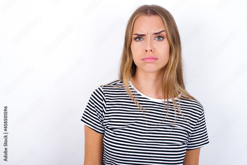 Displeased beautiful blonde girl wearing striped t-shirt on white background frowns face feels unhappy has some problems. Negative emotions and feelings concept Displeased beautiful blonde girl wearing striped t-shirt on white background frowns face feels unhappy has some problems. Negative emotions and feelings concept