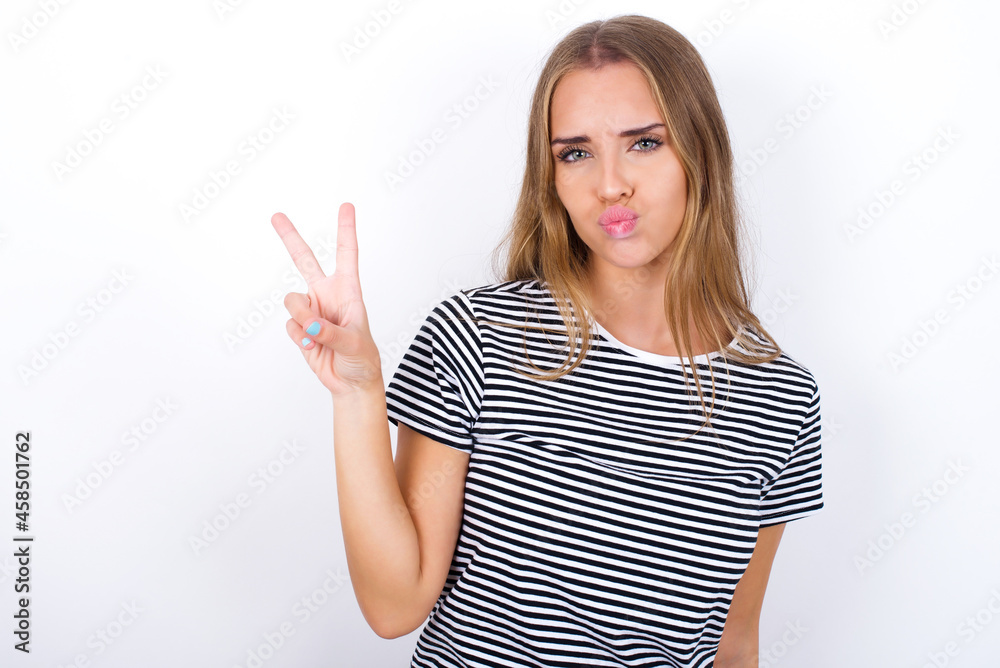 beautiful blonde girl wearing striped t-shirt on white background makes peace gesture keeps lips folded shows v sign. Body language concept beautiful blonde girl wearing striped t-shirt on white background makes peace gesture keeps lips folded shows v sign. Body language concept