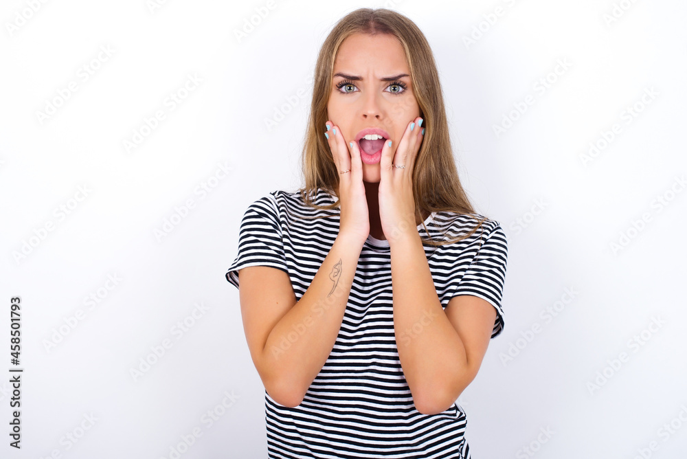 Upset beautiful blonde girl wearing striped t-shirt on white background touching face with two hands