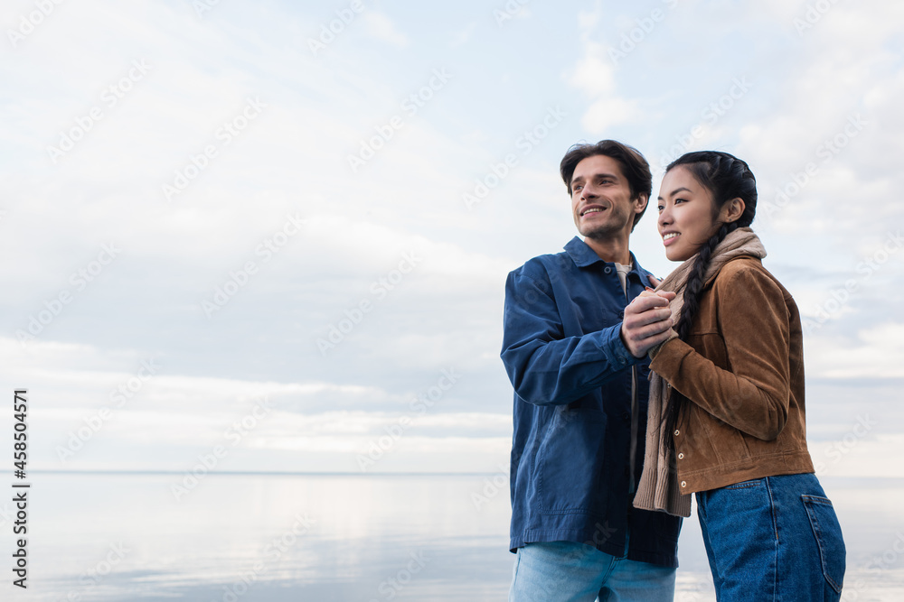 Smiling multiethnic couple holding hands near lake and sky at background Smiling multiethnic couple holding hands near lake and sky at background