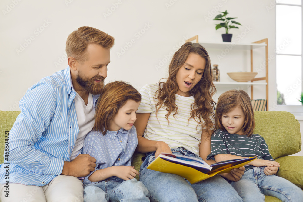 Happy family spending time together. Parents and kids sitting on sofa and reading a book. Beautiful young mother reading interesting stories to her children while sitting on couch at home