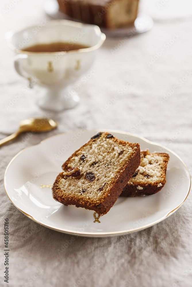 Slice of Pound Loaf Cake with raisins on white porcelain plate and cup of tea on greige linen tablecloth. Selective focus Slice of Pound Loaf Cake with raisins on white porcelain plate and cup of tea on greige linen tablecloth. Selective focus