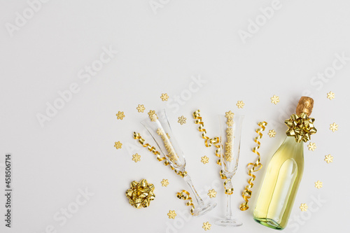 Festive white background with gold decoration , bottle of sparkling wine with two crystal glasses, shiny golden serpentine confetti and glittering snowflakes, golden star, copy space, top view