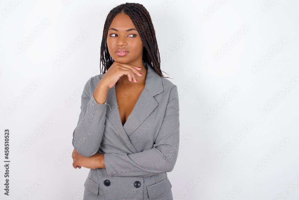Dreamy Young African American businesswoman with braids on white wall with pleasant expression, looks sideways, keeps hand under chin, thinks about something pleasant. Dreamy Young African American businesswoman with braids on white wall with pleasant expression, looks sideways, keeps hand under chin, thinks about something pleasant.