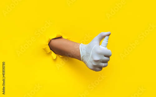 A right doctor's hand in a white medical glove holds a spray sanitizer. Torn hole in yellow paper, copy of the space. Concept of protection against coronavirus, covid-19 and other bacteria and viruses