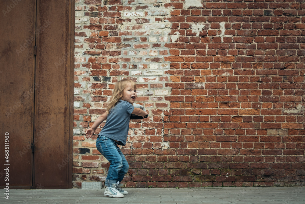 Little cute girl in a blue t-shirt and jeans with a cap is dancing on the background of an old brick wall.