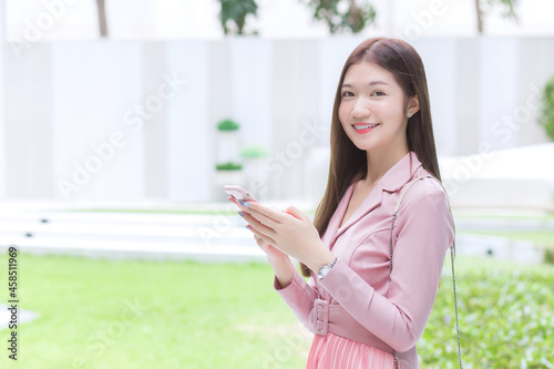 Young Asian professional working woman in pink dress suit holds and looks at her smartphone happily in green natural background.