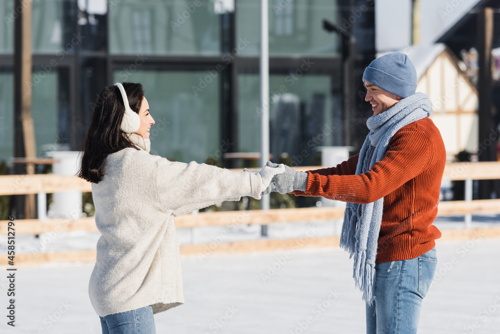 side view of positive young woman in ear muffs holding hands with boyfriend on ice rink