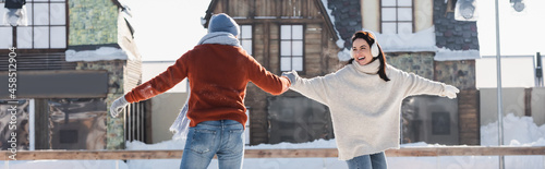 positive young woman in ear muffs holding hands with boyfriend on ice rink, banner