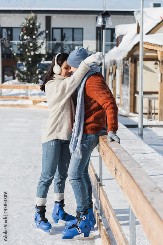 full length of happy woman in ear muffs hugging man in winter hat leaning on wooden border on ice rink