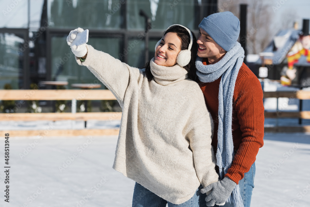happy woman in sweater and ear muffs taking selfie with boyfriend on ice rink