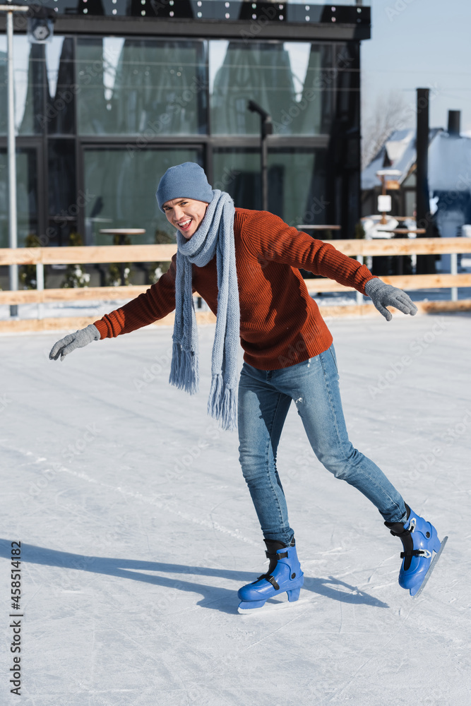 full length of happy young man in sweater, scarf and winter hat skating on ice rink full length of happy young man in sweater, scarf and winter hat skating on ice rink