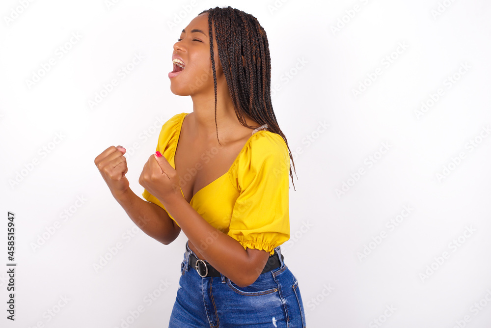 afro american woman with braids wearing sexy yellow t-shirt on white wall excited and glad to achieve victory, clenches fists, screams in excitement with closed eyes,successful person. afro american woman with braids wearing sexy yellow t-shirt on white wall excited and glad to achieve victory, clenches fists, screams in excitement with closed eyes,successful person.