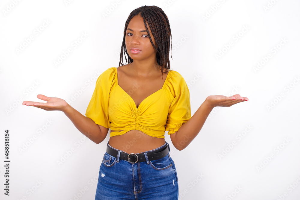 afro american woman with braids wearing sexy yellow t-shirt on white wall looks uncertain shrugs shoulders.