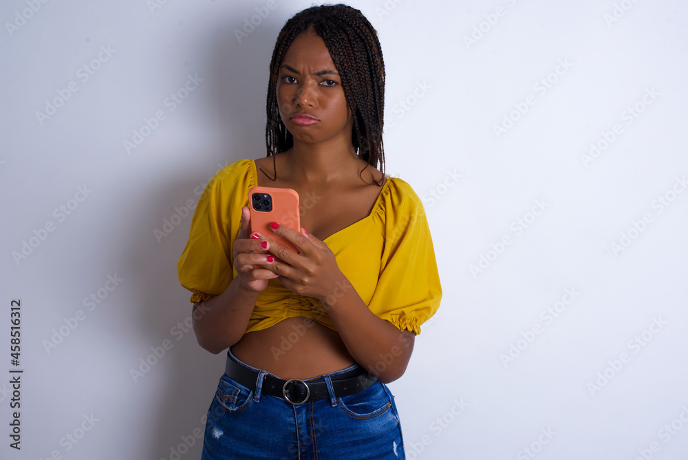 Portrait of a confused afro american woman with braids wearing sexy yellow t-shirt holding mobile phone and shrugging shoulders and frowning face. Portrait of a confused afro american woman with braids wearing sexy yellow t-shirt holding mobile phone and shrugging shoulders and frowning face.