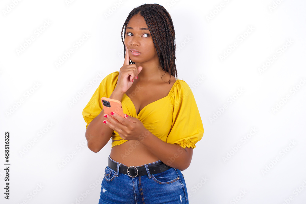 Image of a thinking dreaming afro american woman with braids wearing sexy yellow t-shirt on white wall using mobile phone and holding hand on face. Taking decisions and social media concept. Image of a thinking dreaming afro american woman with braids wearing sexy yellow t-shirt on white wall using mobile phone and holding hand on face. Taking decisions and social media concept.
