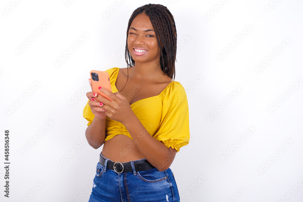 Pleased afro american woman with braids wearing sexy yellow t-shirt on white wall using self phone and looking and winking at the camera. Flirt and coquettish concept. Pleased afro american woman with braids wearing sexy yellow t-shirt on white wall using self phone and looking and winking at the camera. Flirt and coquettish concept.