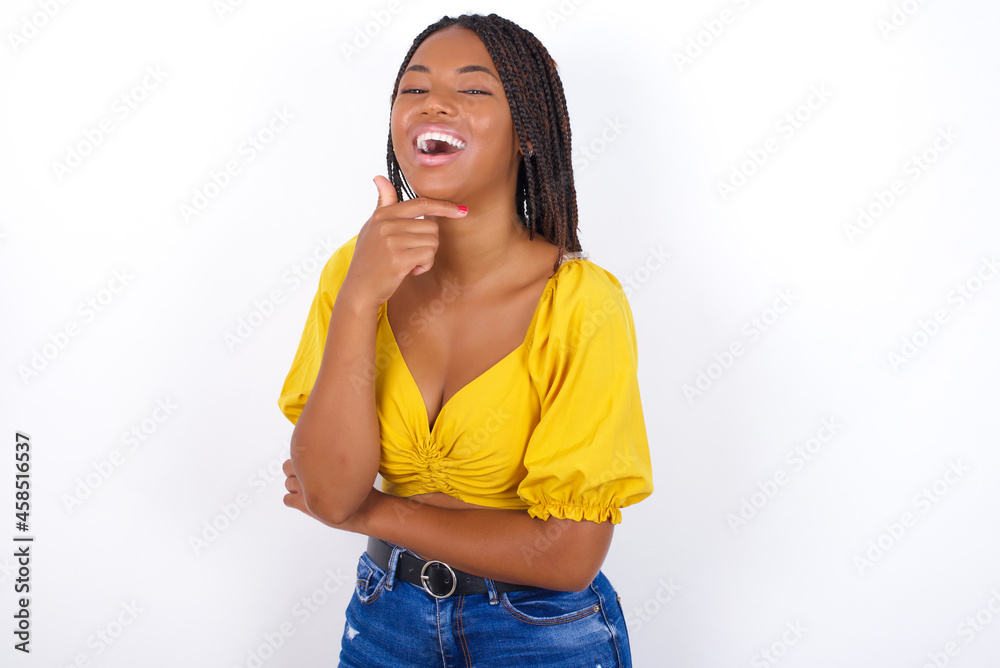 afro american woman with braids wearing sexy yellow t-shirt on white wall laughs happily keeps hand on chin expresses positive emotions smiles broadly has carefree expression