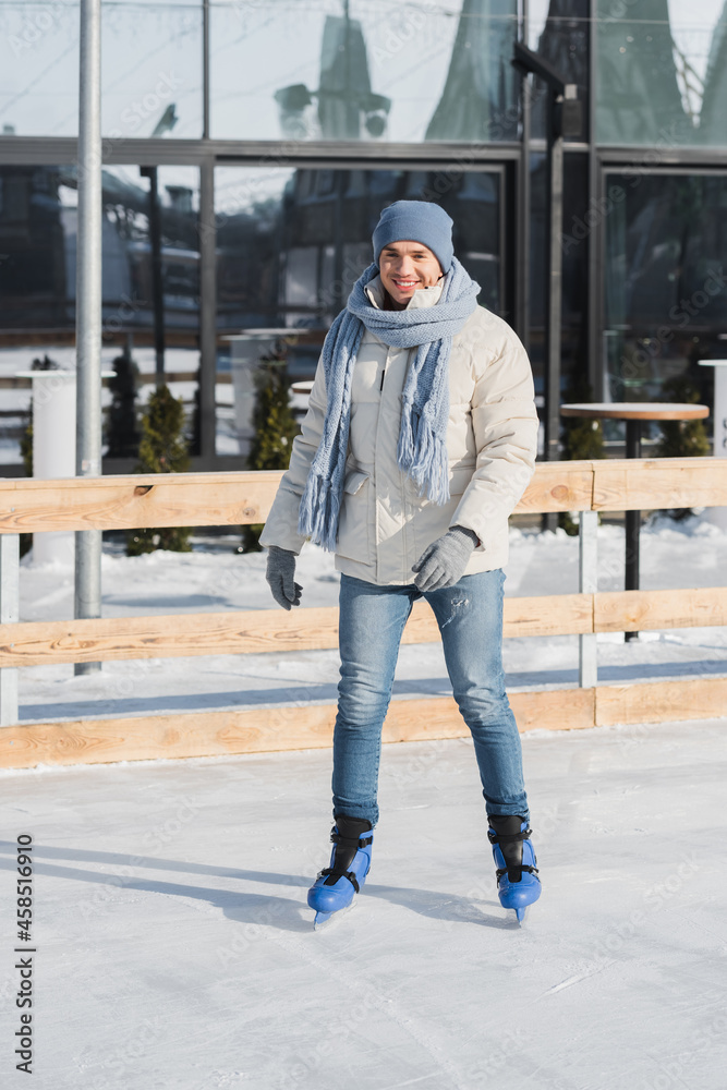 full length of happy young man in scarf, winter hat and ice skates on ice rink full length of happy young man in scarf, winter hat and ice skates on ice rink