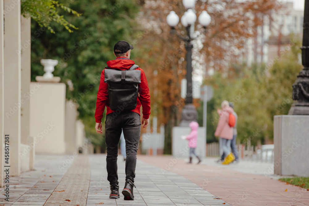 Back view man in red jacket with backpack walking along autumn alley Back view man in red jacket with backpack walking along autumn alley