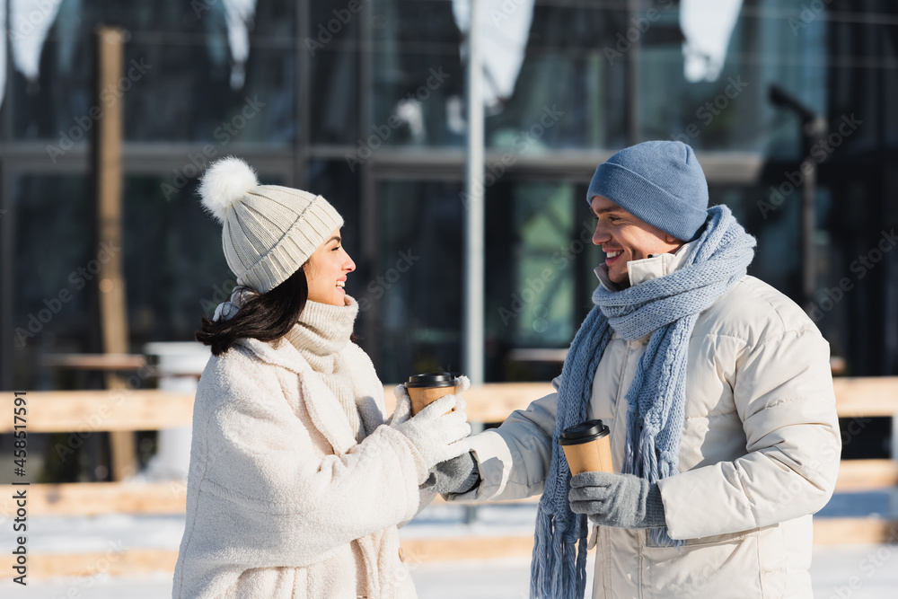 happy young man giving paper cup to smiling girlfriend in winter hat