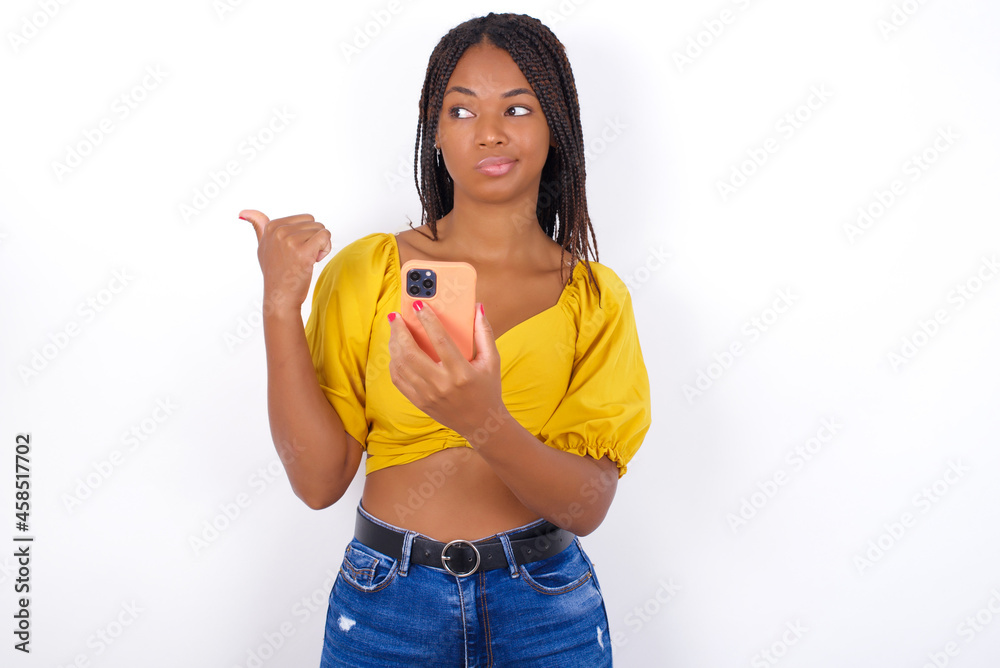 afro american woman with braids wearing sexy yellow t-shirt on white wall points thumb away and shows blank space aside, holds mobile phone for sending text messages. afro american woman with braids wearing sexy yellow t-shirt on white wall points thumb away and shows blank space aside, holds mobile phone for sending text messages.