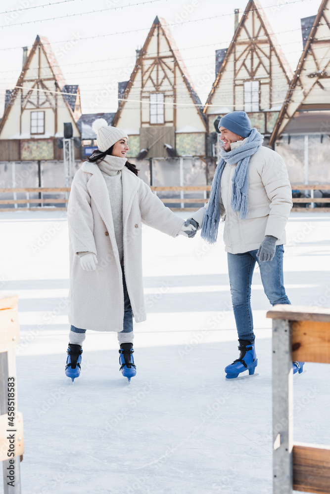 full length of happy young man in winter hat and ice skates holding hands with smiling girlfriend on ice rink