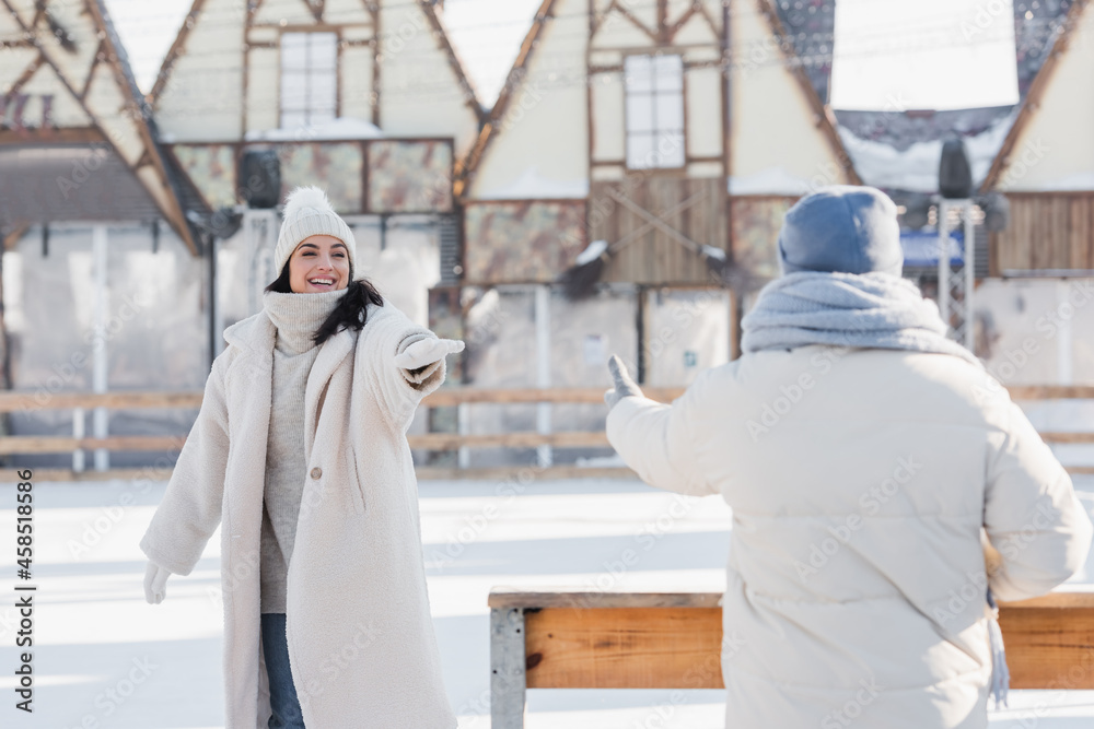 smiling young woman in winter hat standing with outstretched hand near blurred boyfriend