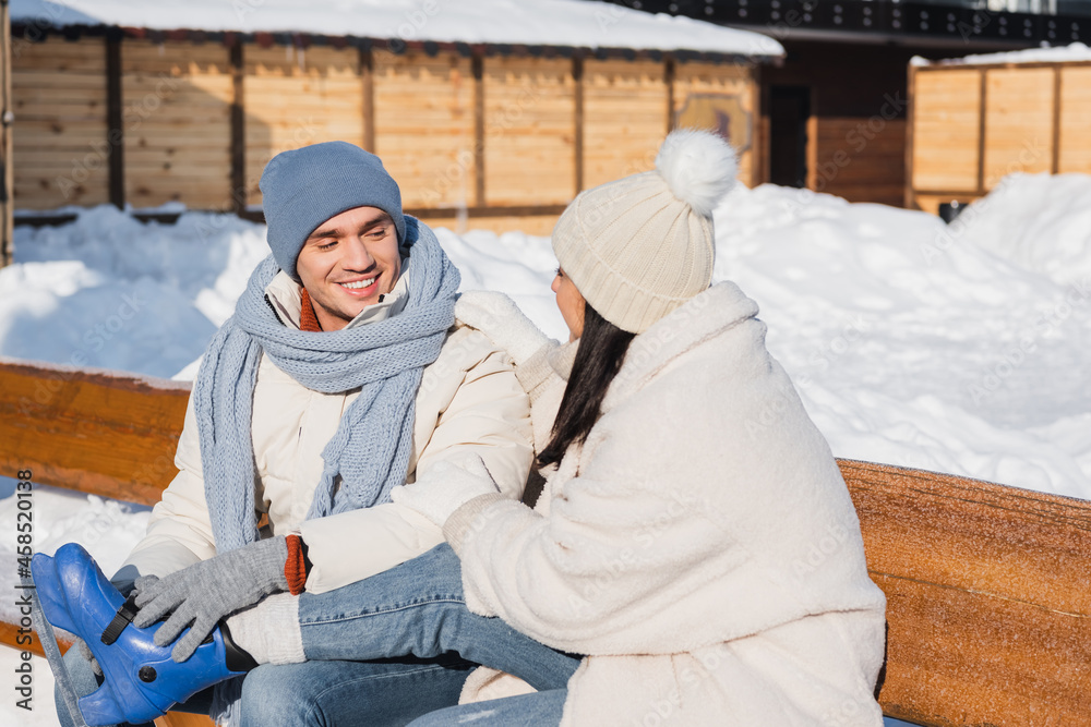 happy young man in winter hat sitting on bench and wearing ice skates on girlfriend happy young man in winter hat sitting on bench and wearing ice skates on girlfriend