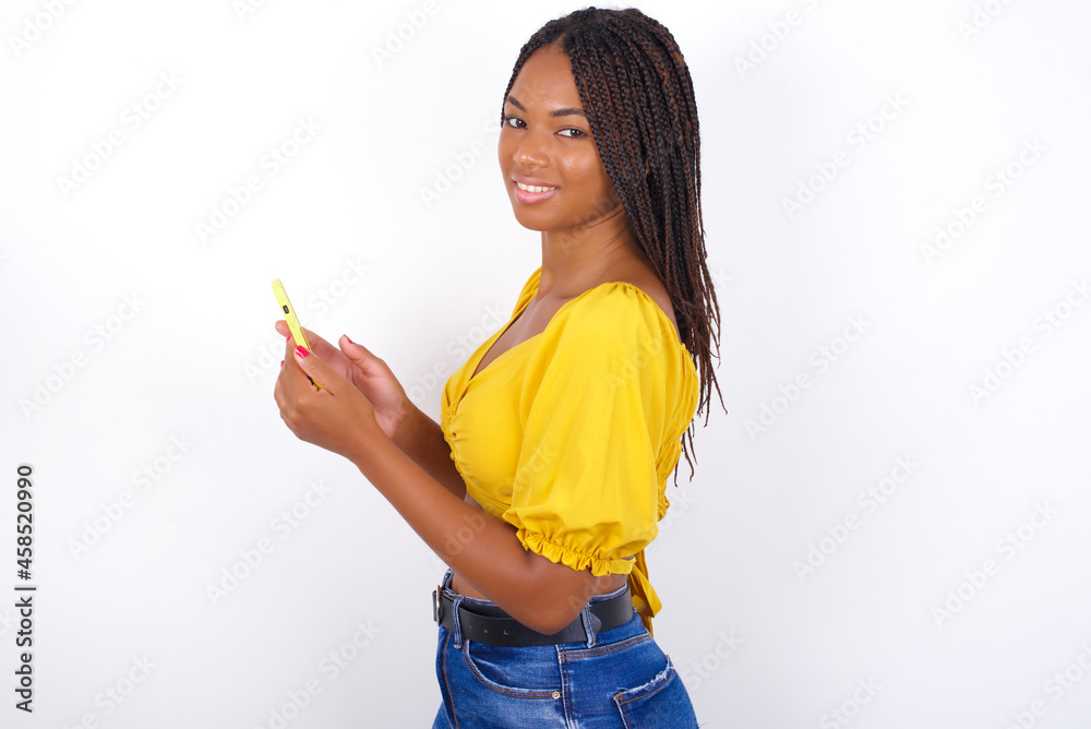 Rear view photo portrait of afro american woman with braids wearing sexy yellow t-shirt on white wall using smartphone smiling Rear view photo portrait of afro american woman with braids wearing sexy yellow t-shirt on white wall using smartphone smiling