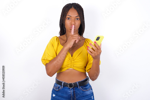 afro american woman with braids wearing sexy yellow t-shirt on white wall holding modern gadget ask not tell secrets