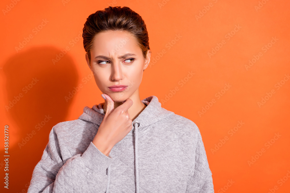 Photo of pretty unsure young woman dressed grey hoodie arm chin looking empty space isolated orange color background