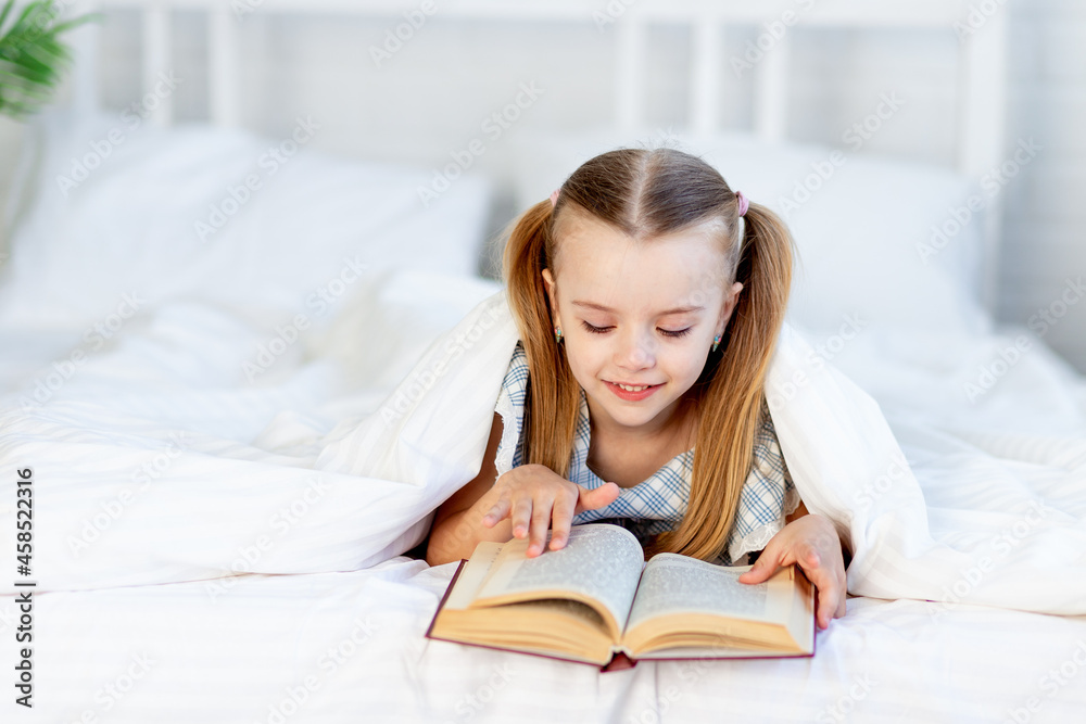 a girl child is reading a book on the bed at home on a white cotton bed under a blanket and smiles sweetly a girl child is reading a book on the bed at home on a white cotton bed under a blanket and smiles sweetly