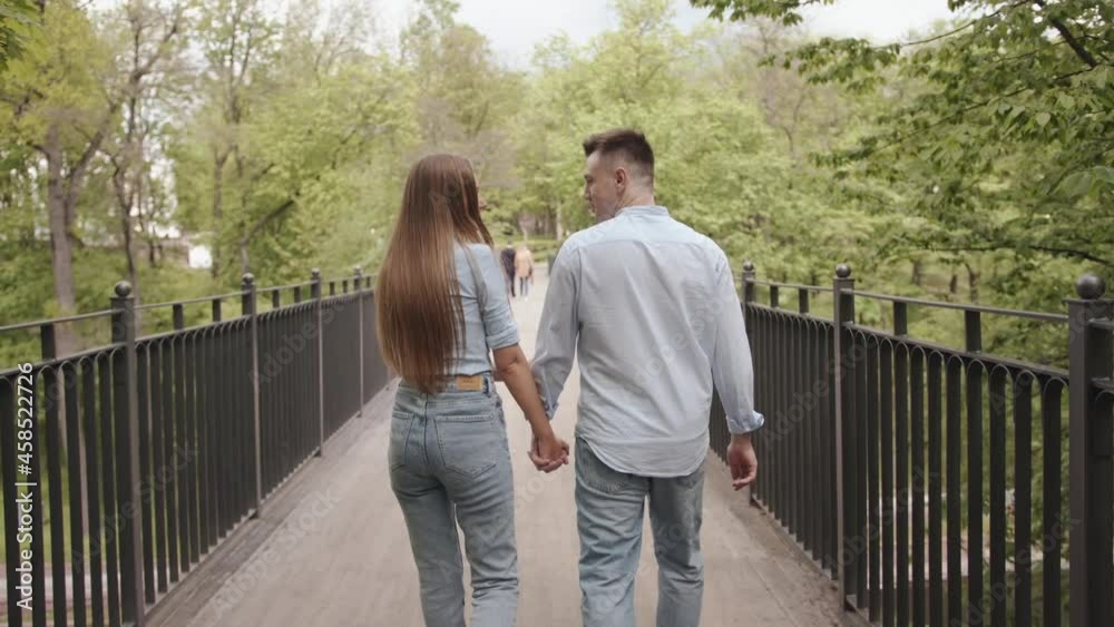 A young couple in love goes along the bridge over the river in the park on a summer day and smiling looking at each other. Slow motion A young couple in love goes along the bridge over the river in the park on a summer day and smiling looking at each other. Slow motion