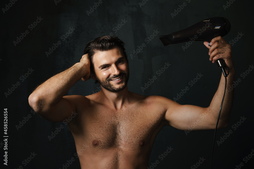 Young half-naked man smiling while using hair dryer