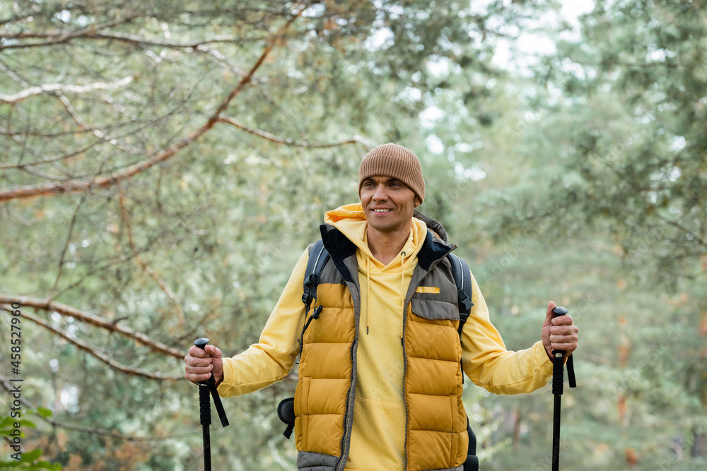 joyful traveler with trekking poles looking away while standing in forest