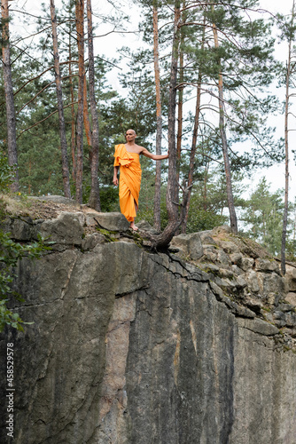 full length view of buddhist monk in orange kasaya walking on rocky cliff in forest photo