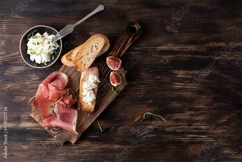 Cutting board with prosciutto, slices of bread and ricotta on a dark wooden background, italian ham with figs.