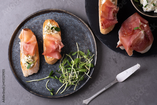 Italian appetizer of bread and prosciutto, crostini with ham, ricotta and microgreens on a gray background.