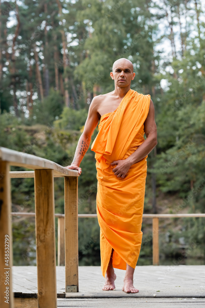 full length view of buddhist monk standing near wooden fence in forest - obrazy, fototapety, plakaty 